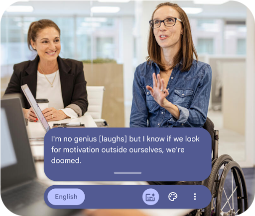 Two women having a conversation in the workplace. One is seated in a wheelchair and wears glasses. The other sits in a chair with a smile.