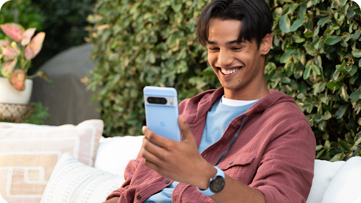A man sitting on a patio outdoors while looking at a smartphone and smiling.