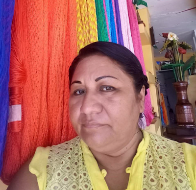 A portrait of Irene, a woman wearing a yellow sleveless top, gazing at the camera against a rainbow backdrop of woven textiles.